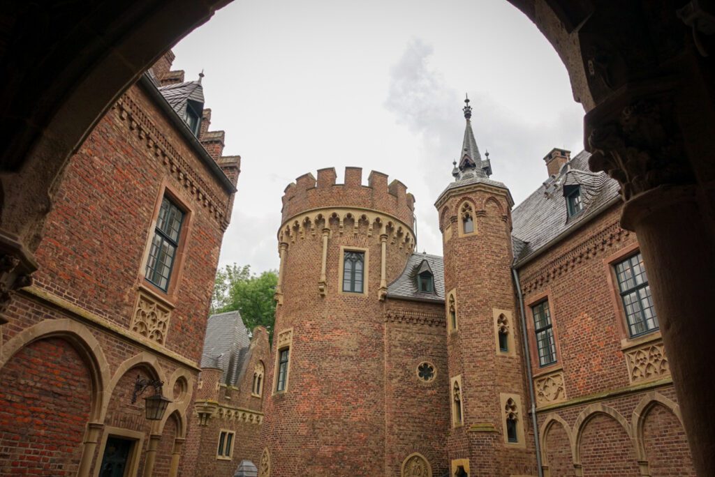 A picture of Schloss Paffendorf, near to Cologne. The picture is taken through one of the arches of the castle and the the turrets, spires and orange-red brick walls and detailing of the castle contrast to the grey clouds in the sky. Paffendorf belongs to the hidden gems surrounding Cologne and the most fairytale-like secret castles in Germany