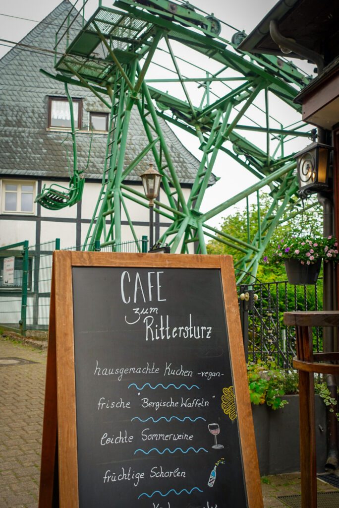 A picture of the oldest double seater cable car in Germany, with a green metal structure. In front of the cable car is a sign for 'Cafe zum Rittersturz', advertising homemade cakes and Bergische Waffeln. Not only is Schloss Burg one of the most historic secret castles in Germany, but the cable car is one of the most unqiue too