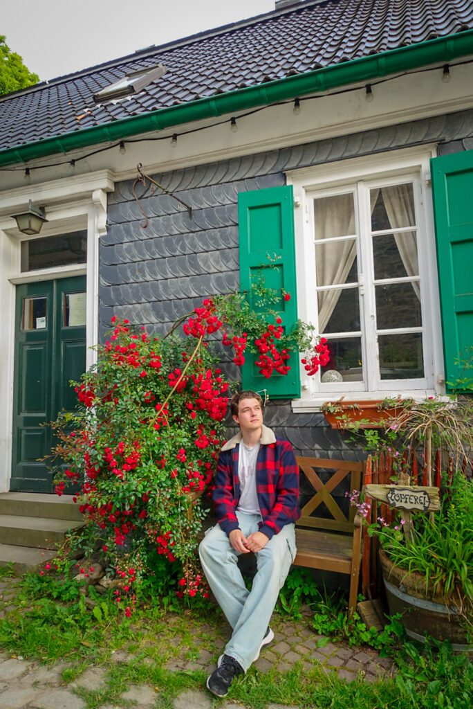 A picture of myself sitting on a bench, in a red checkered jacket and jeans, looking out from a bench at Schloss Burg in Solingen. Behind me is part of the castle and next to me a red flower bush.