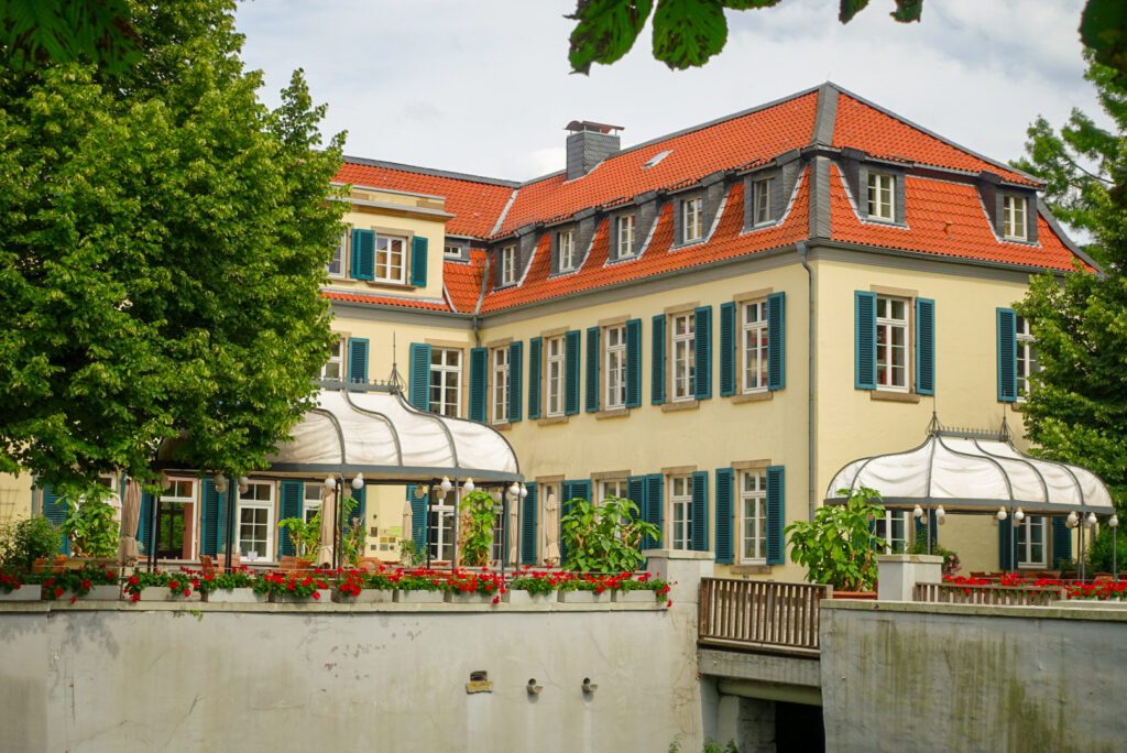 A picture of Schloss Berge in Gelsenkirchen. The red tiled roof of the hotel contrasts to the cream coloured walls and the white windows are decorated with dark blue shutters. There are white canopies on the front of the castle and the bridge is decorated with bright red flowers.