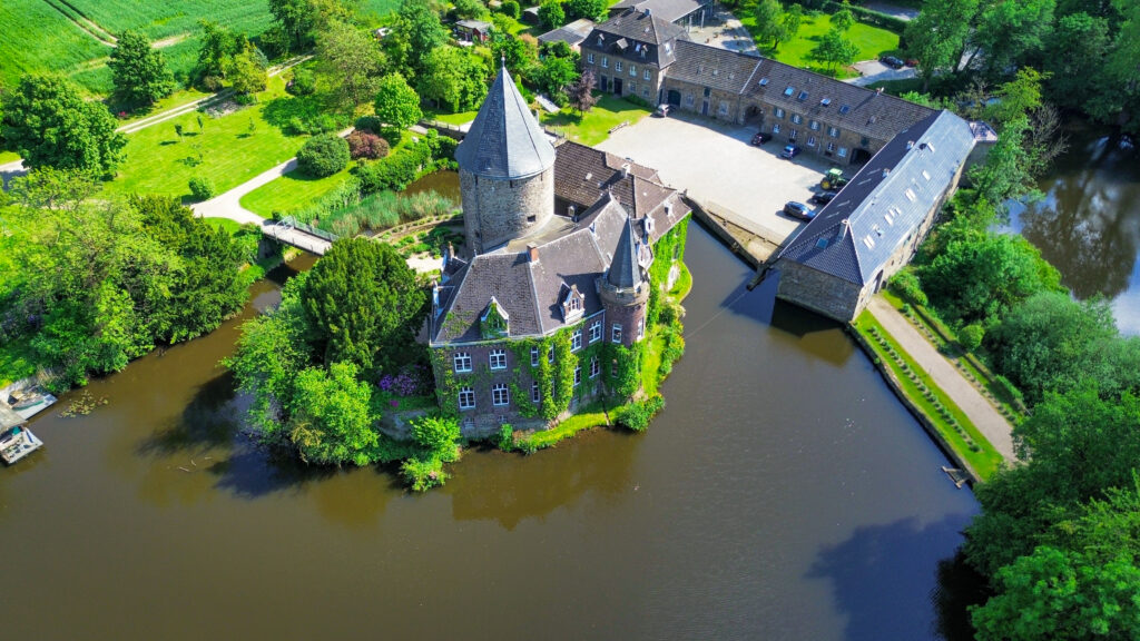 A picture of Schloss Linnep near Düsseldorf- a hidden European travel destination. The stone castle is surrounded by a moat and ivy dresses the brickwork of the castle looking out towards the lake.