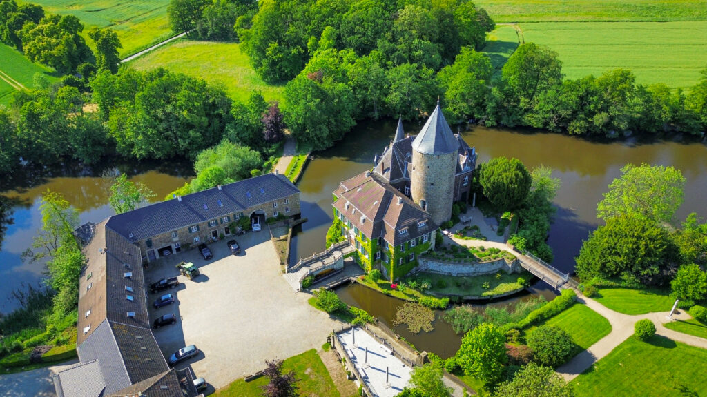 A picture of Schloss Linnep in Düsseldorf from above. Multiple cars are parked on the courtyard of the castle and the pointed defensive tower is surrounded by green gardens and also a lake.