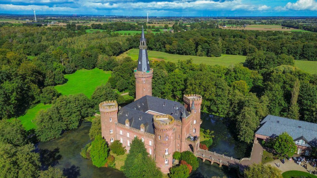 An aerial photo of Schloss Moyland. The castle has four spires with red brick, the roof has grey tiles and there is one large spire with a silver coloured turret. The castle is surrounded by a moat, green and orange trees and wind turbines in the distance.