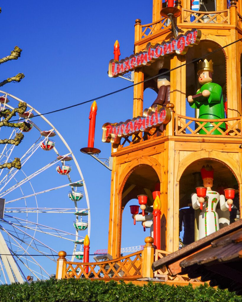 A picture of a Christmas pyramid and the ferris wheel at the Luxembourg Christmas markets. There are various christmas figures inside the pyramid, which is adorned with candles on the outside.