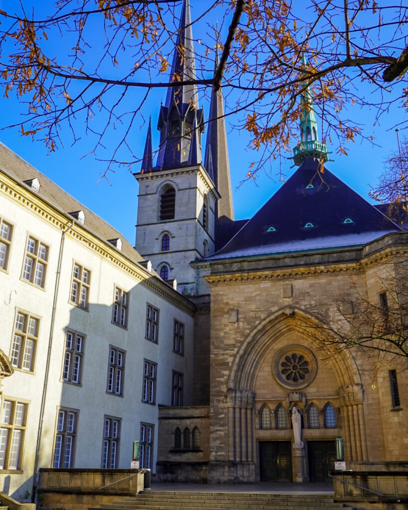 A picture of the Notre-Dame cathedral in Luxembourg. Travel to Luxembourg with the deutschlandticket using the RE11 from Koblenz. The cathedral dates back to the 15th century, has sand-coloured stone against a blue sky and autumn leaves are hanging in-front of the spire.