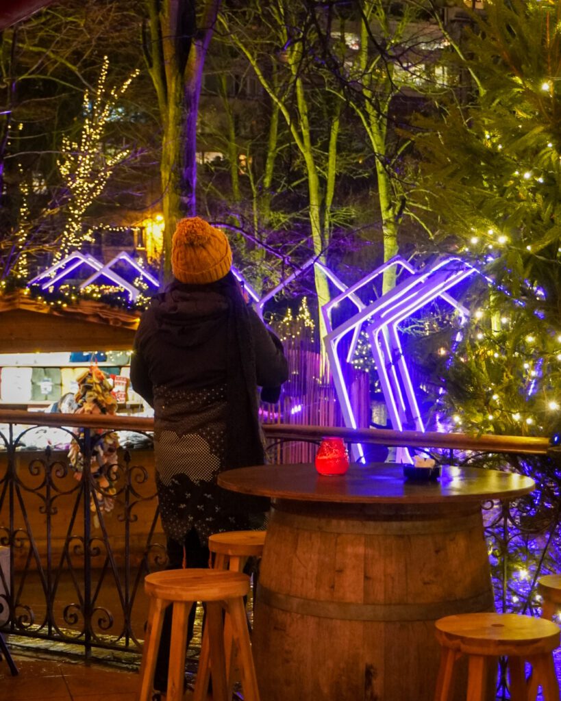 A picture of the christmas markets in Ostende- just 15 minutes away from the city of Bruges and an alternative winter destination. You can see a person standing with their back to the camera, taking a photo of the light arches through the trees.
