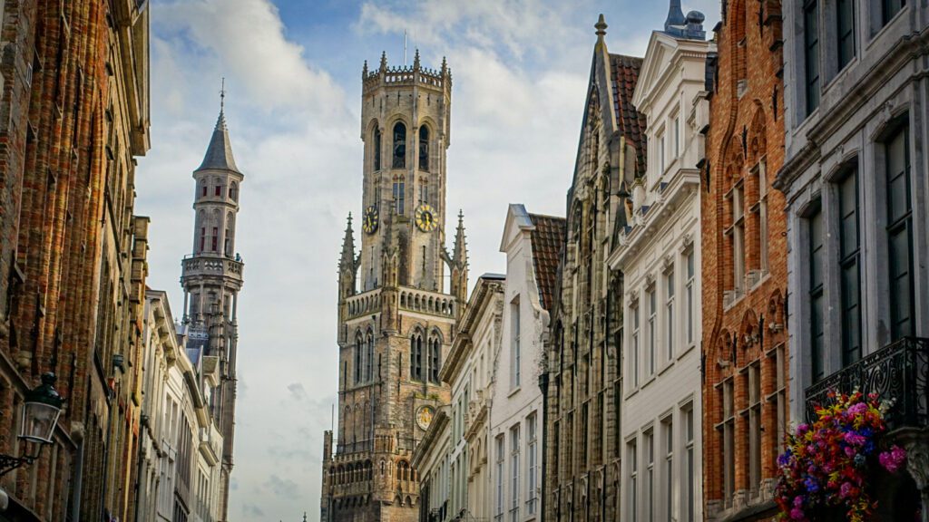 A landscape view of the fairy-tale facades in Bruges, Belgium. In the distance you can see blue sky amongst white floating clouds. The 83meter high Belfort tower is in the centre of the picture in the distance. On the tower you can see a clock and a viewing platform, which provides views onto the whole of Bruges
