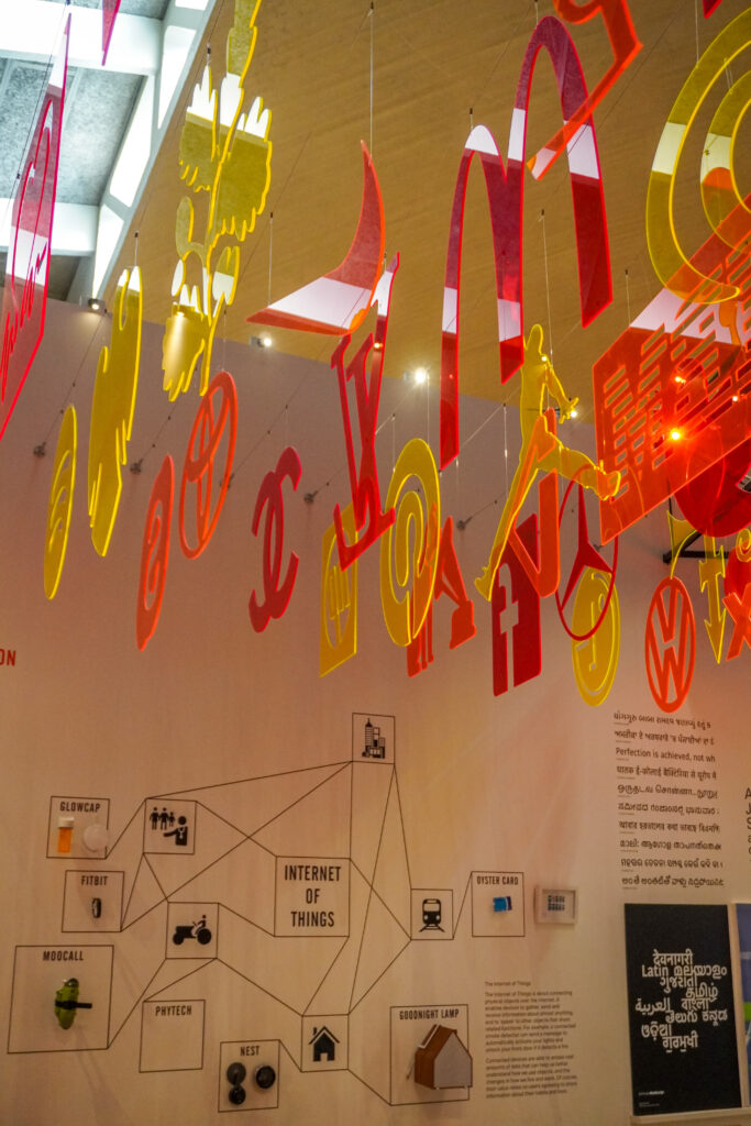 Acrylic neon lettering hangs down from the ceiling in the permanent collection of the Design Museum. The permanent collection is called 'Designer, Maker, User'. The lettering hanging down from the ceiling are yellow, orange and red in colour.