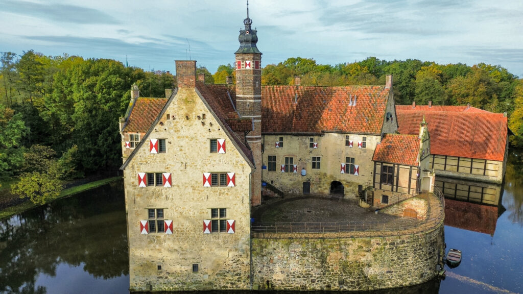 Vischering castle dates back to the 13th century and is surrounded by a moat. The stone walls of the castle are lined with red and white shutters, and the red brick roof stands out in the surrounding backdrop of autumn leaves.