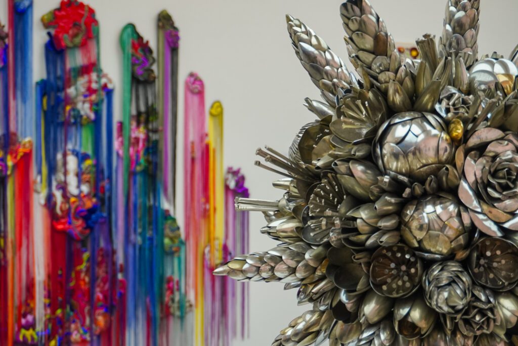 A silver aluminium sculpture is placed in front of a colourful wall display at the Saatchi gallery- the wall display is multi-coloured like a rainbow.