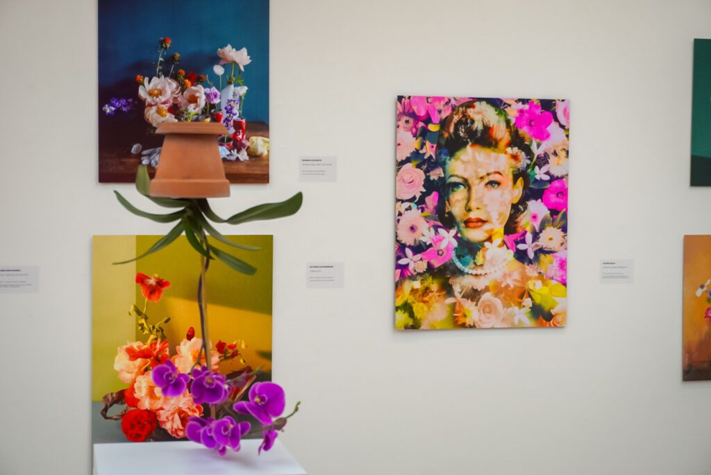 Contemporary art lines the walls of the flowers exhibition at the Saatchi Gallery. An orchid is suspended upside down, on a white pillar. The orchid has purple petals and a brown vase, which is inverted.