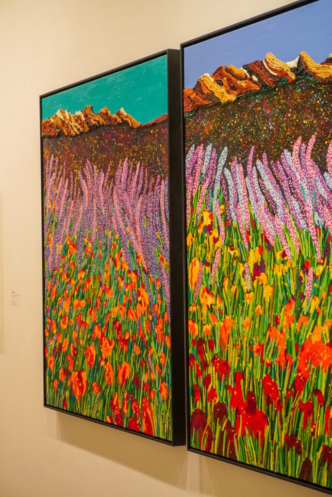 Two paintings are hung on one of the walls of the double-floor gallery. The paintings are identical and depict a mixture of colours. Along with a sea of colourful flowers- from red through to purple, there is a mountain backdrop against a blue sky.