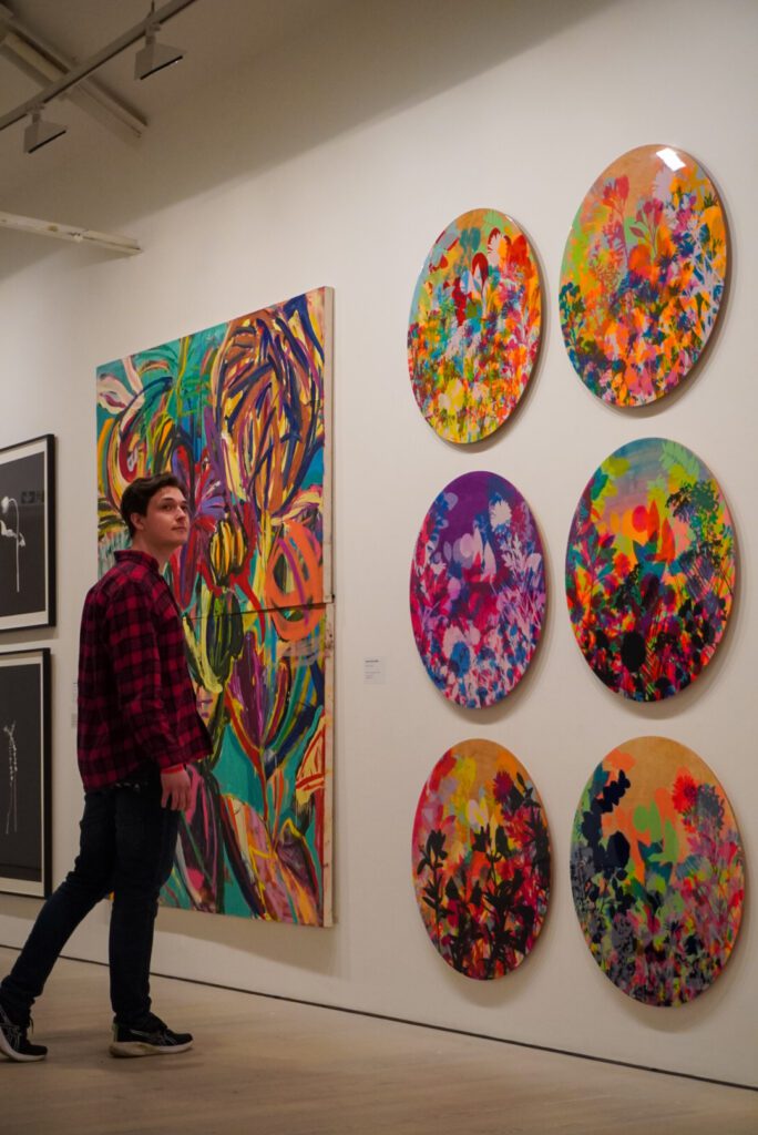 Six round paintings with different colours are mounted on the wall. The round paintings are made out of a multitude of colours, from green and orange to a rich purple. I - Traveller James- am standing in a red checked shirt and blue jeans in front of the paintings, looking over my shoulder and into the disttance.