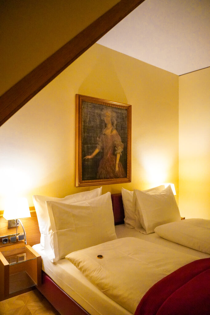 The bedrooms of the De Medici Living Hotel are decorated with cream coloured walls, oil paintings on the wall and the double bed is made up with two pillows on each side, duvets and a red sheet. There is a chocolate on each side of the bed as a welcoming gift.