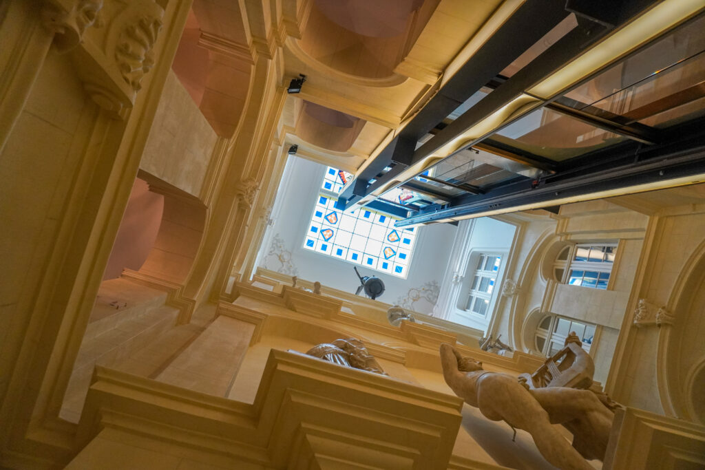 In the Living Hotel De Medici in Düsseldorf you will discover a secret panoramic elevator. The black track of the elevator extends towards the ceiling, which is made out of a glass roofing with mosaic tiling. At the top of the elevator on the third floor is a bronze sculpture, depicting a roman Godess.