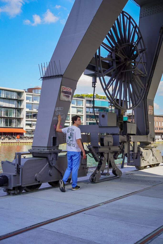 The city harbour is known for its industrial architecture and incredible views onto the canal. Myself, Traveller James, can be seen in this photo, with a white t shirt and blue trousers, walking underneath one of the industrial cranes at the harbour in Muenster, which would have been previously used for marine-trade. On the other side of the canal you can see flats, which look out onto the water as well as restaurants.