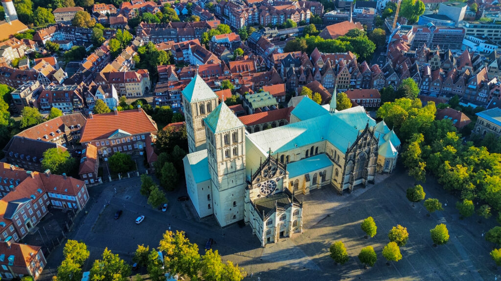 Domplatz in Muenster from a bird's eye perspective- the city's cathedral is surrounded by trees in their autumn colours: green, orange and golden yellow. The cathedral has a turquoise roof and there are views onto the cities red-roof housing in the background as well as to the Prinzipalmarkt on the right hand side of the photo