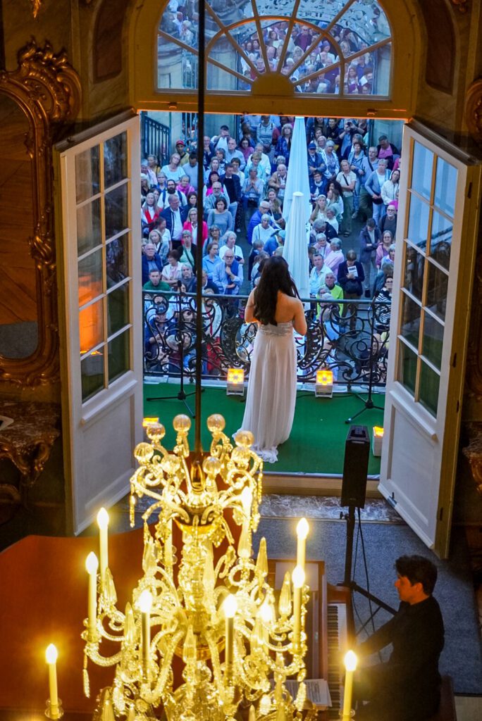 The Schauraum takes place once yearly in the city of Muenster, during which you can experience a free opera concert. The singer is standing in a white dress on the balcony of the building and on the inside there is a piano-player, who is playing the background music. The room is crowned by a chandelier, which is located above the piano player.